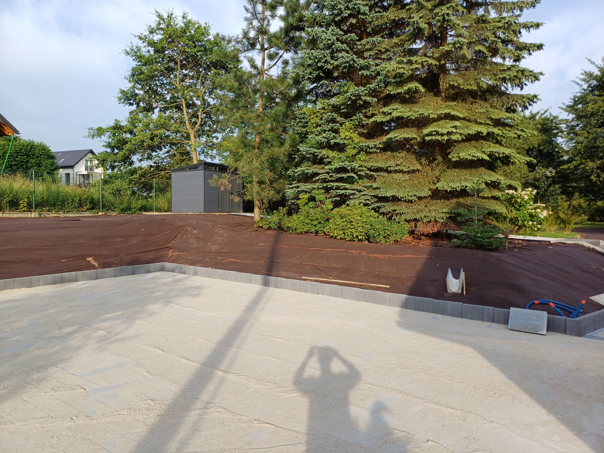 Przygotowany teren pod kostkę brukową z widoczną podsypką piaskową, obrzeżami z kostki i brązową włókniną, w tle szary blaszany domek narzędziowy i zieleń ogrodu.