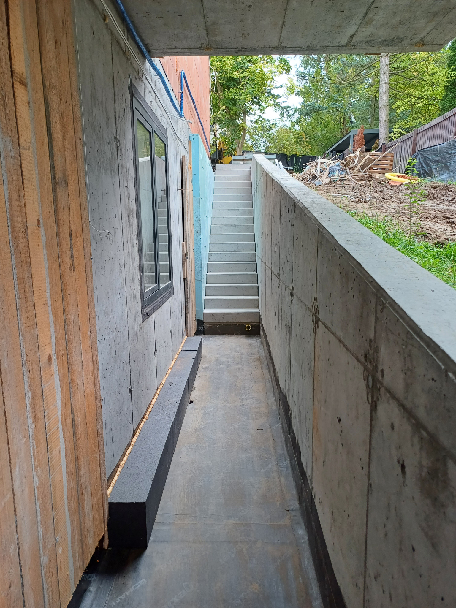 Betonowy wykop pod schody zewnętrzne z oknem w ścianie i widoczną instalacją.