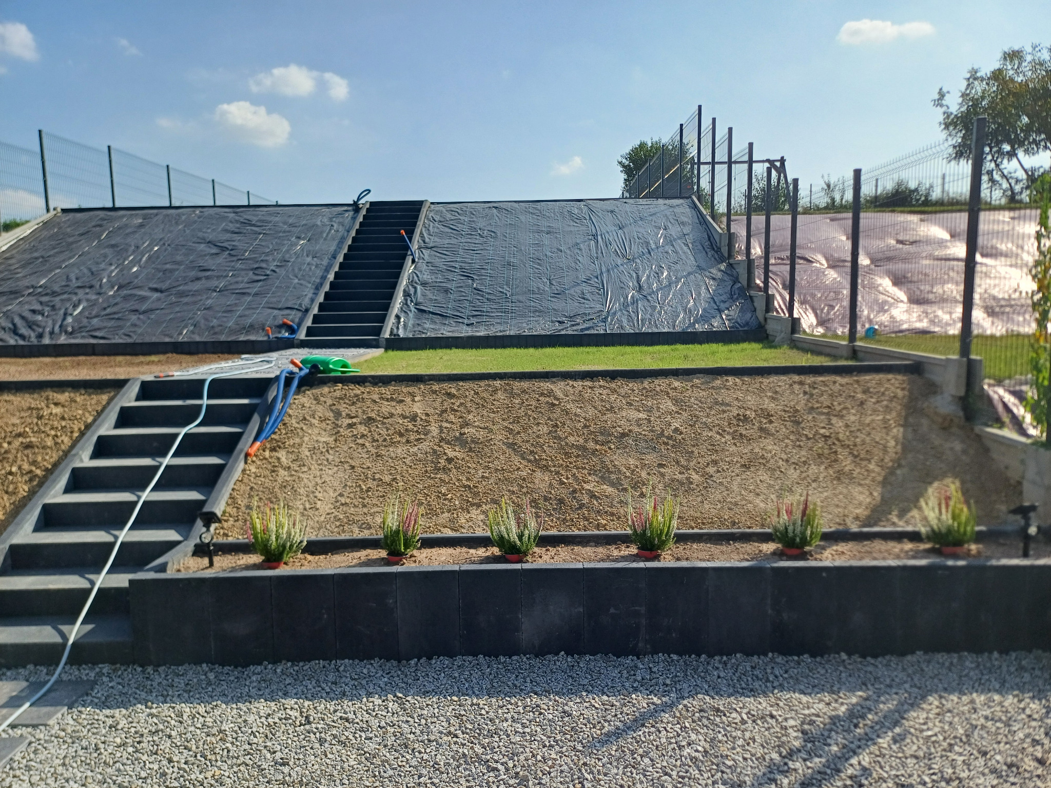 Przygotowanie terenu pod ogród: czarna folia na skarpie, schody z kostki, obsypany piaskiem taras, obrzeże z doniczkami i wysypany żwirem teren przed domem.