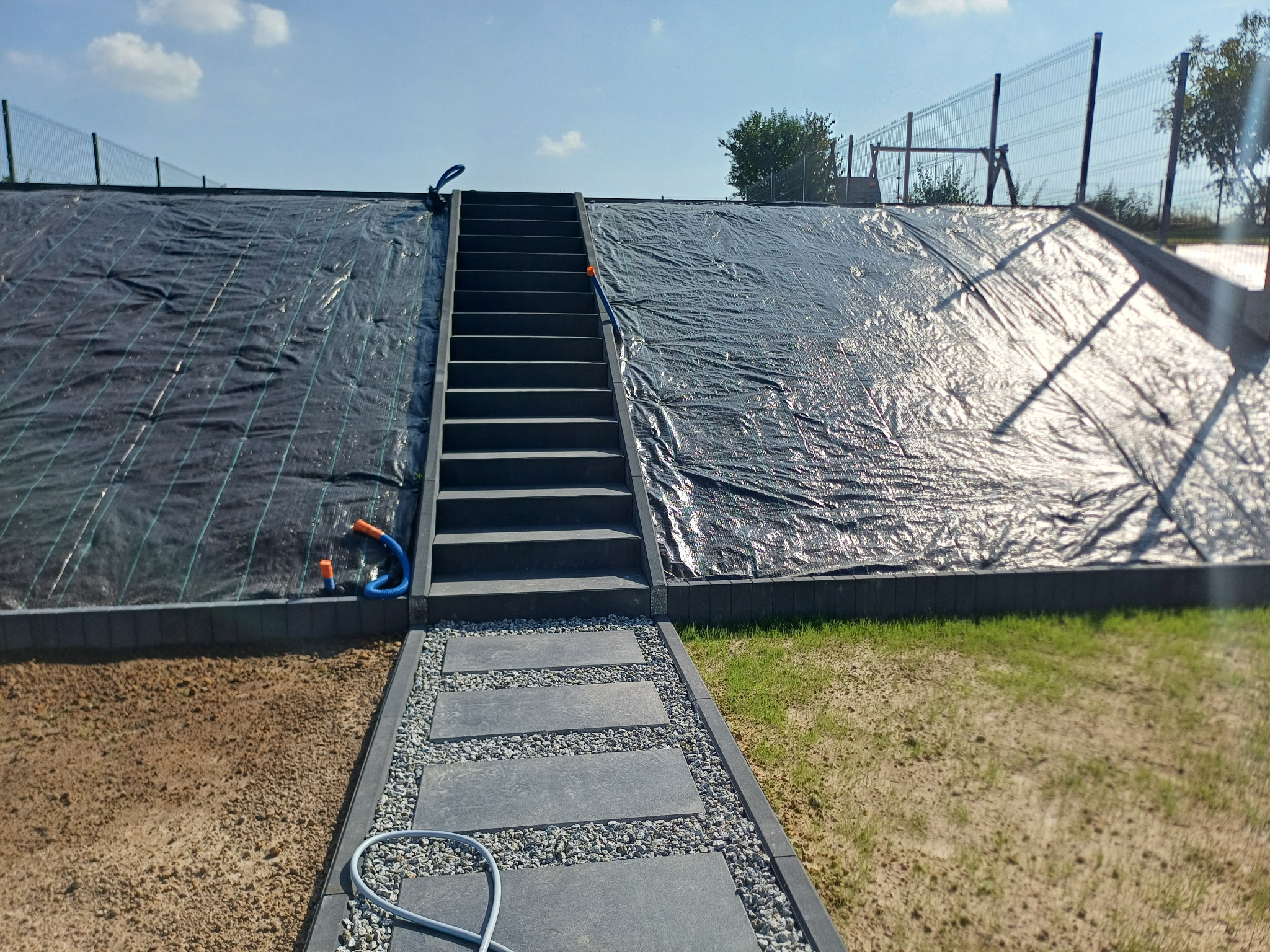 Ułożona ścieżka z płyt betonowych prowadząca do schodów na wzmocnionym nasypie, zabezpieczonym czarną agrowłókniną i ogrodzonym siatką.