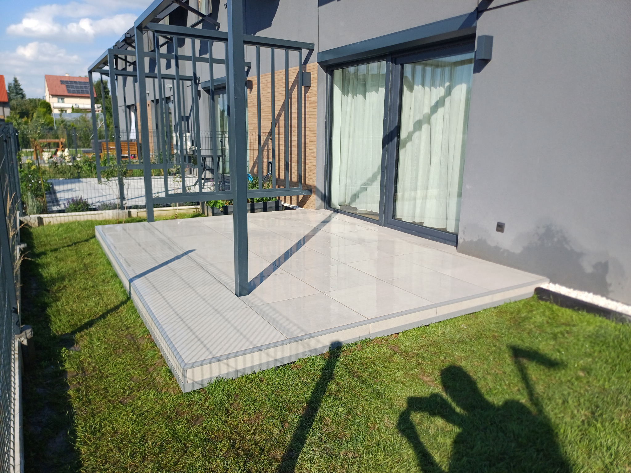 Nowoczesny taras z jasnych płytek ceramicznych, zadaszony metalową konstrukcją, przylegający do szarego budynku z przesuwnymi drzwiami tarasowymi, otoczony trawnikiem.