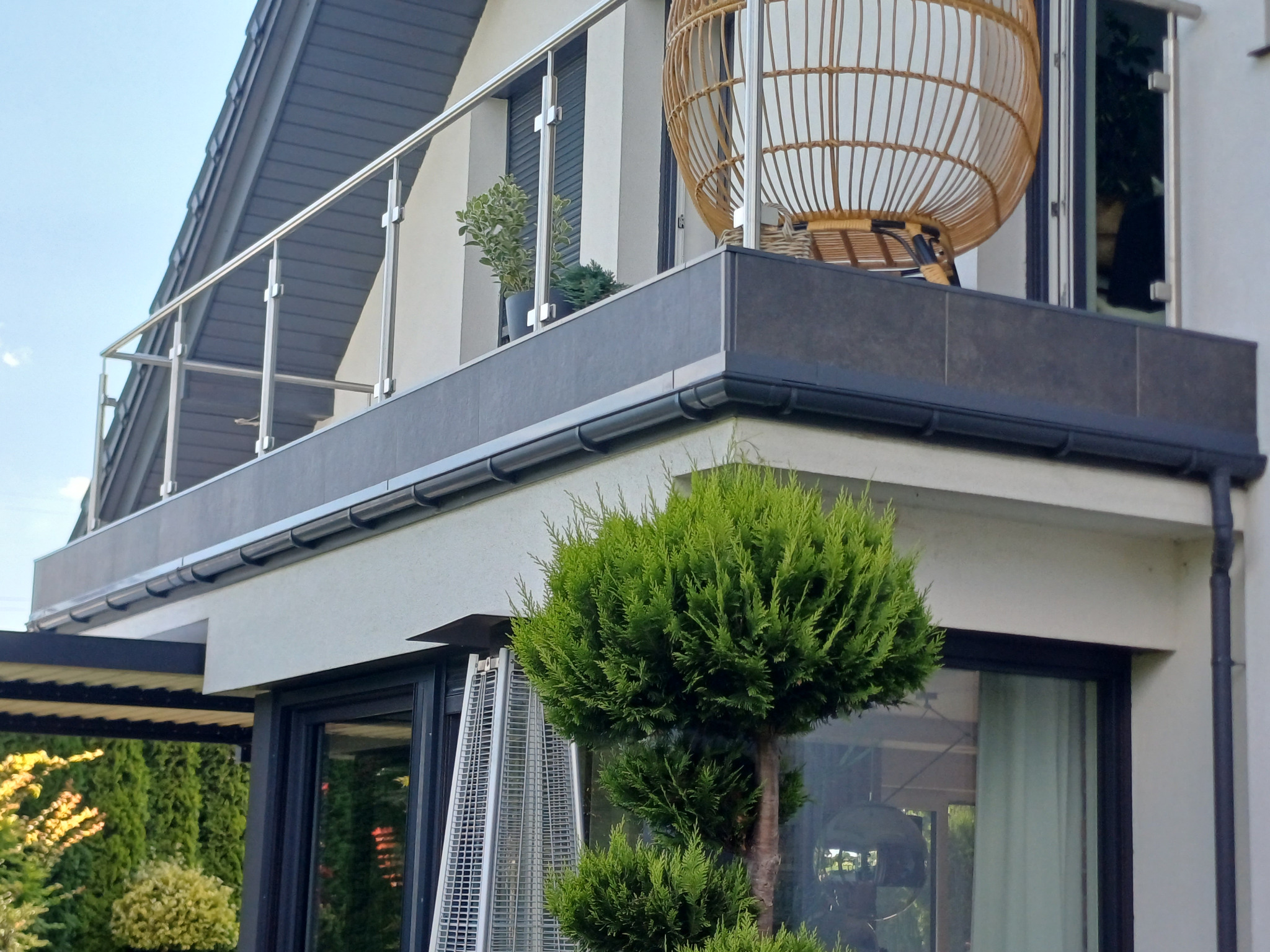 Nowoczesny balkon z szarą płytką, metalową balustradą i wiklinowym fotelem wiszącym, widok z dołu z ozdobnym drzewkiem iglastym na pierwszym planie.