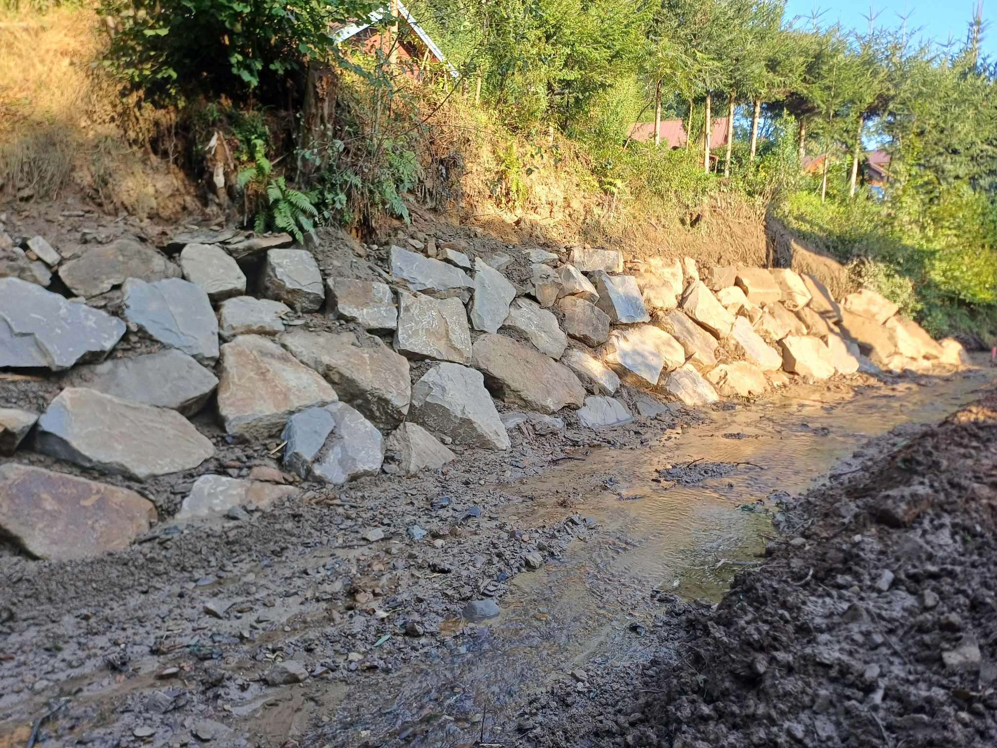 Skarpa zabezpieczona murem oporowym z kamieni, z widocznym ciekiem wodnym w korycie rzeki i ziemnym brzegiem.