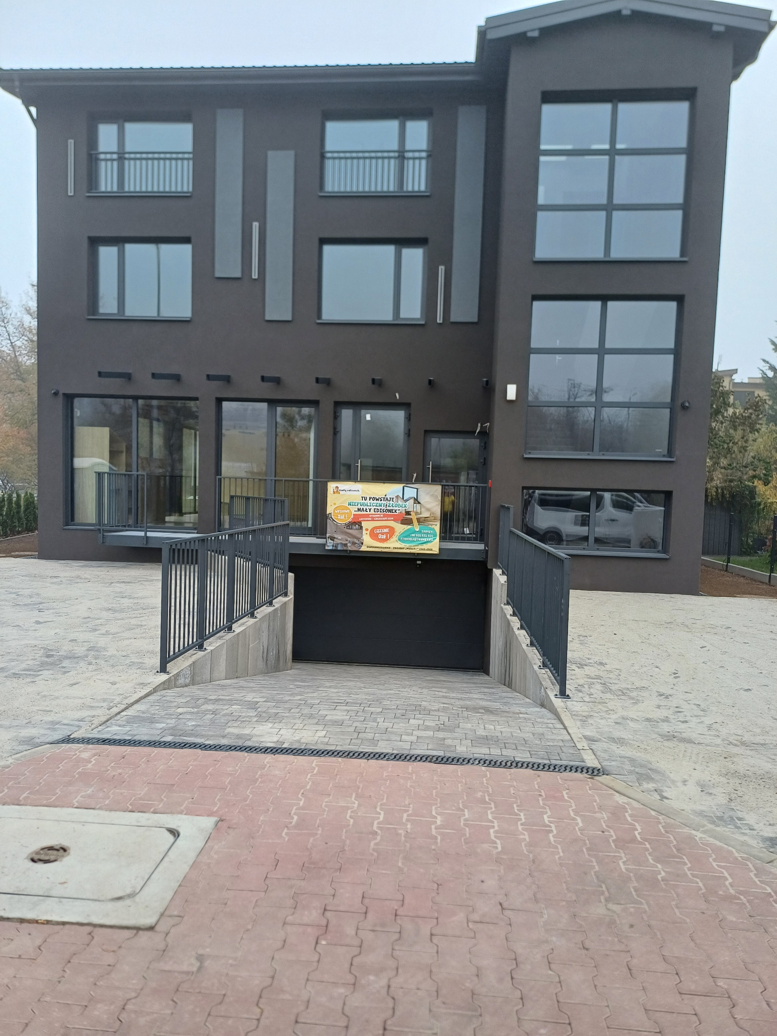 Nowoczesny dom z podjazdem wyłożonym czerwoną kostką brukową przechodzącą w szarą, metalowa barierka, brama garażowa i baner reklamujący żłobek.