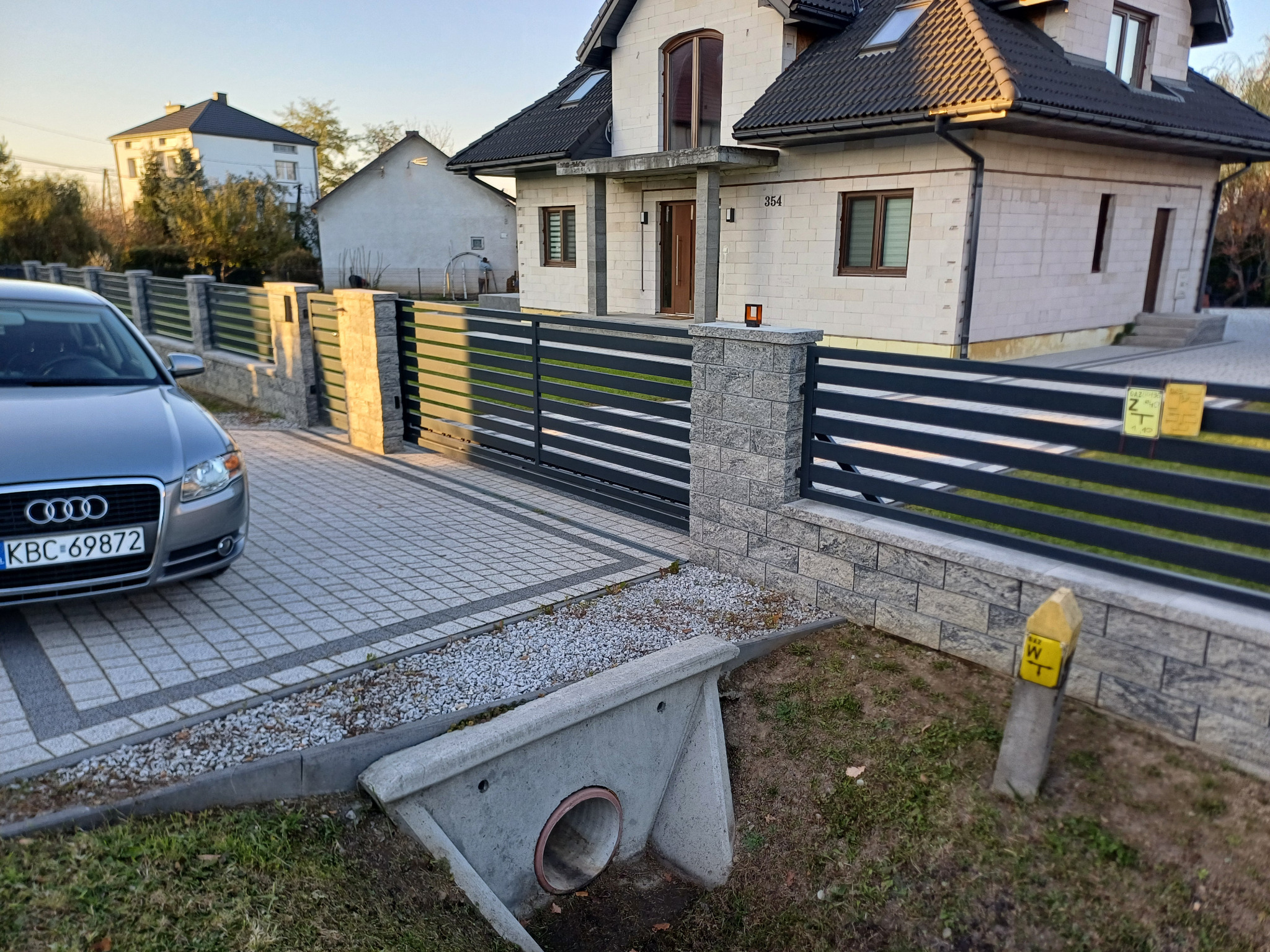 Nowoczesne ogrodzenie metalowe z elementami z szarego kamienia w Dziewinie, widok na posesję z kostką brukową i szarym Audi zaparkowanym obok.