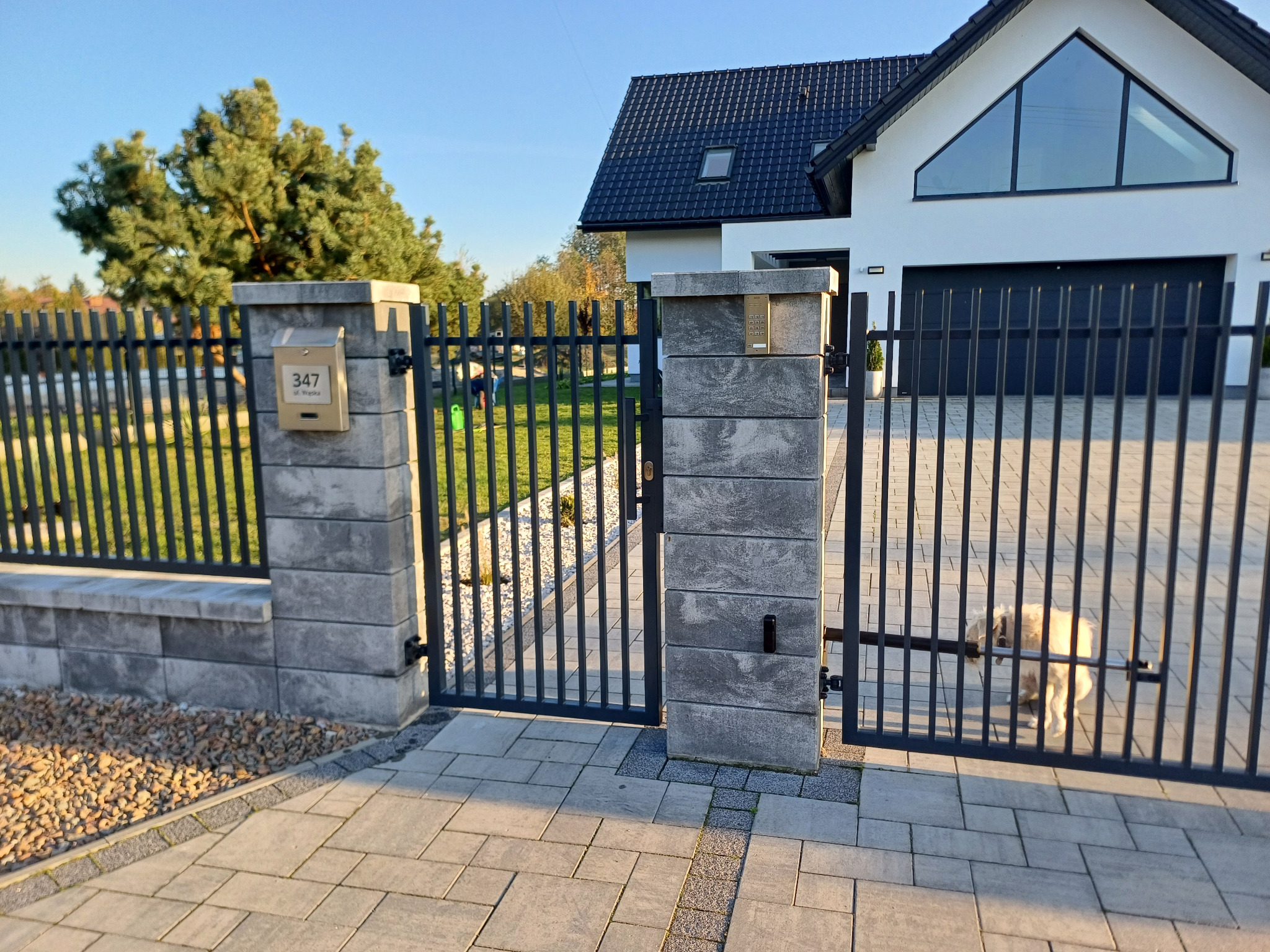 Nowoczesne ogrodzenie metalowe w kolorze grafitowym z murowanymi słupkami z szarej kostki, skrzynka na listy z numerem domu, w tle dom z dużym oknem trójkątnym i kostka brukowa na podjeździe.