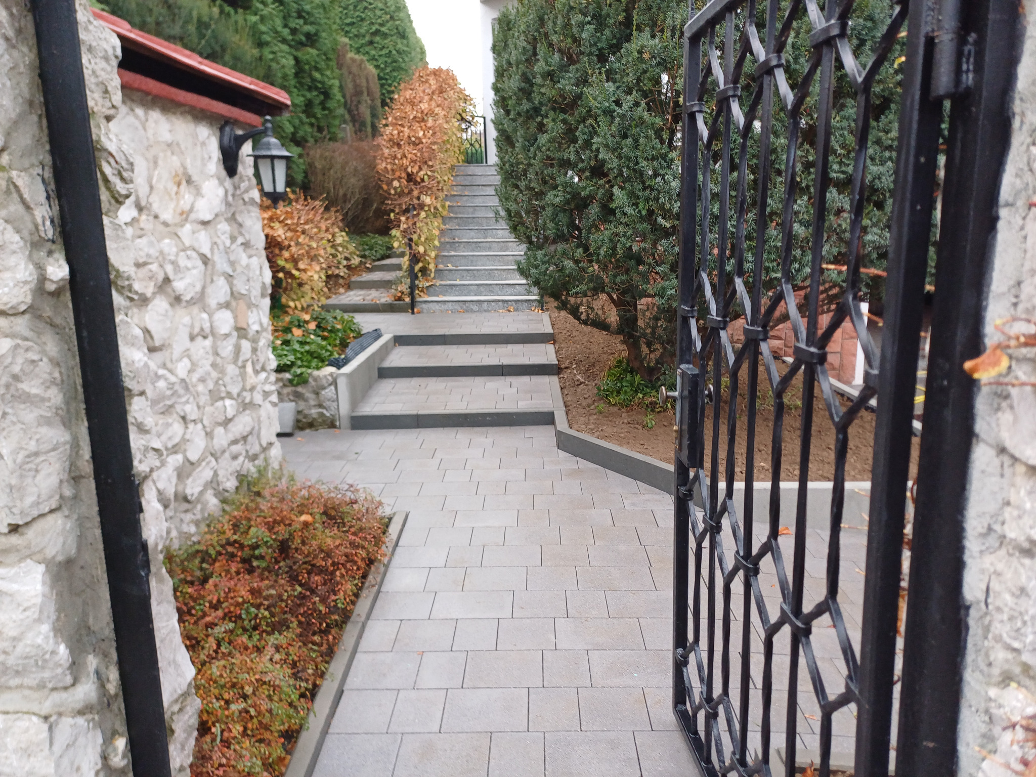 Ułożona z jasnoszarej kostki brukowej ścieżka prowadzi do schodów i otwartej, czarnej, metalowej bramy. Kamienny murek z lampą po lewej stronie, z prawej krzewy.