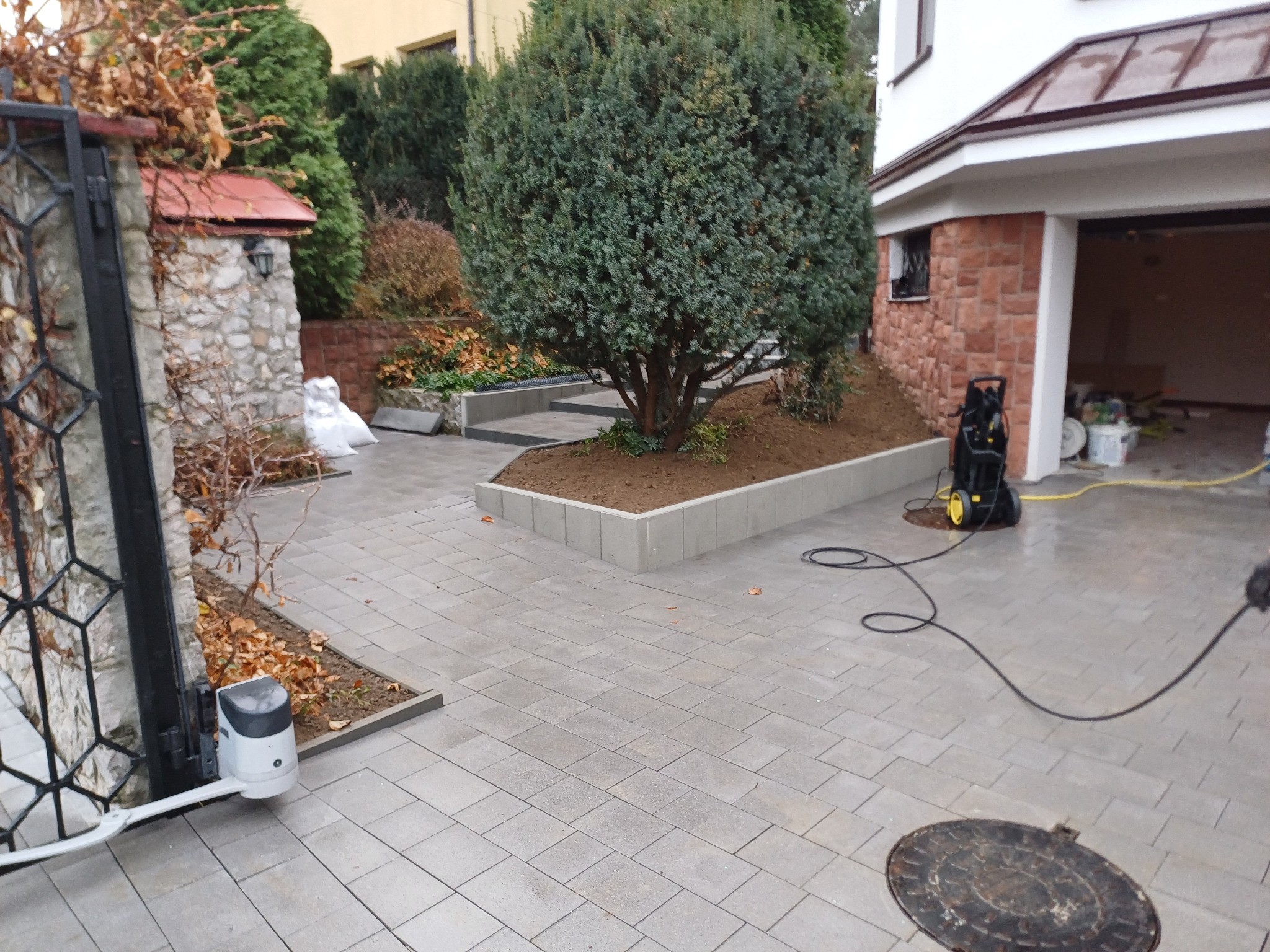 Ułożona szara kostka brukowa na podjeździe z automatyczną bramą w tle, obok rabata z drzewem i budynek z garażem, czyszczona myjką ciśnieniową.