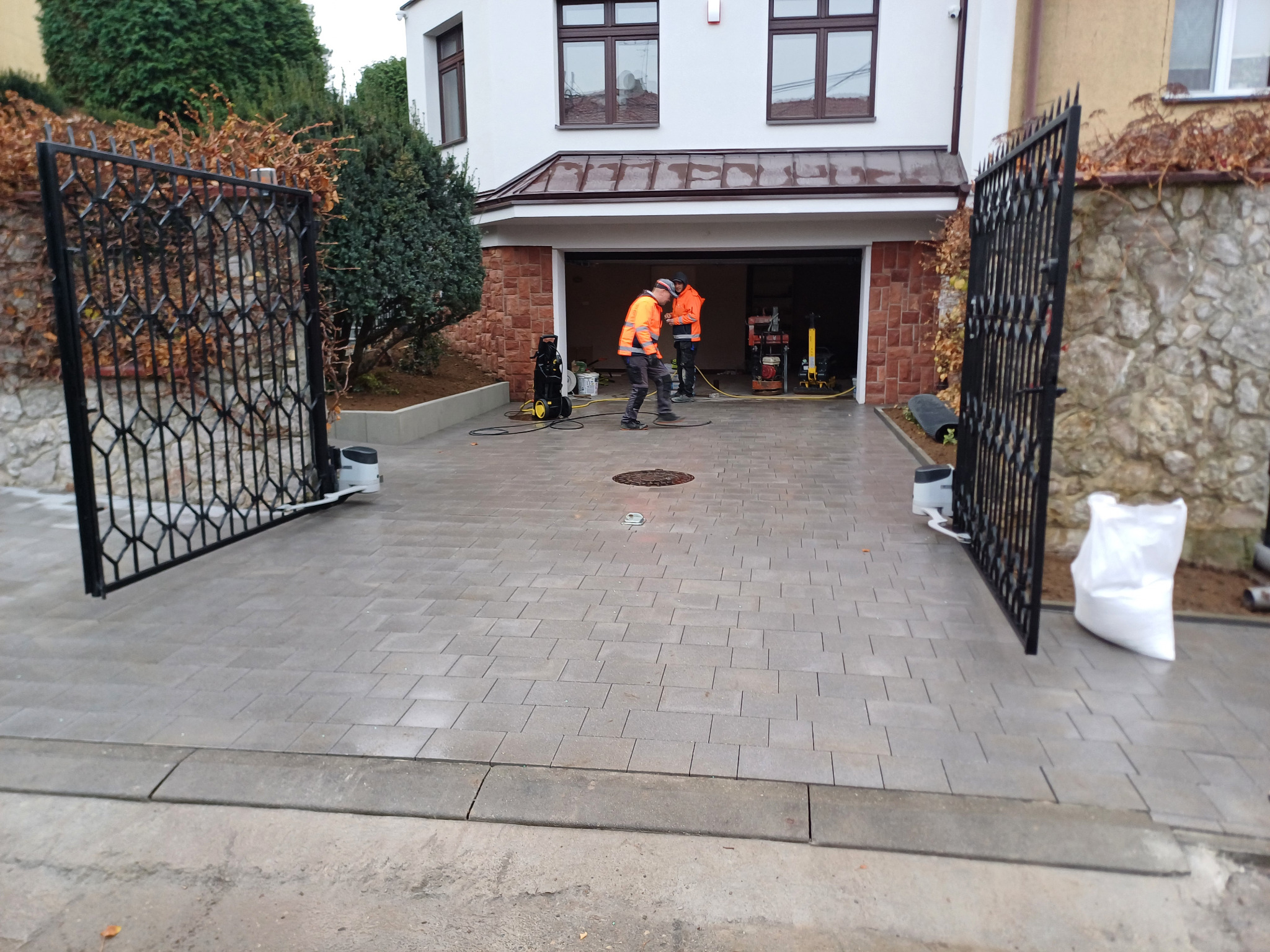 Nowo ułożona, szara kostka brukowa na podjeździe przed domem z otwartą, ozdobną, metalową bramą wjazdową. Dwóch pracowników w pomarańczowych kamizelkach czyści powierzchnię za pomocą myjki...