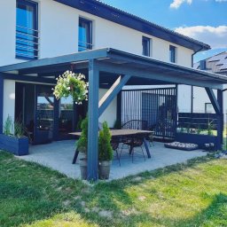 Self Made - Damian Greiss - Nowoczesna, grafitowa konstrukcja zadaszenia tarasu przy białym domu, z wiszącą kompozycją kwiatową i meblami ogrodowymi, na tle błękitnego nieba.