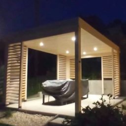 Self Made - Damian Greiss - Oświetlona altana z jasnego drewna z lamelowymi ścianami bocznymi, osłonięty mebel ogrodowy wewnątrz, widok nocny.