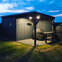 Self Made - Damian Greiss - Nowoczesna, szara konstrukcja z drewna z zadaszeniem, oświetleniem i miejscem na grilla, otoczona trawnikiem i ogrodzeniem panelowym, widziana o zmierzchu.
