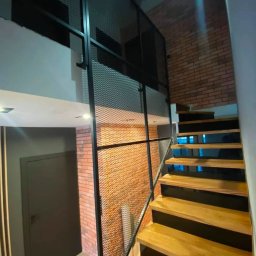 Self Made - Damian Greiss - Schody drewniane z czarnymi podstopnicami i metalową balustradą z siatki, widok z dołu, ceglana ściana w tle.