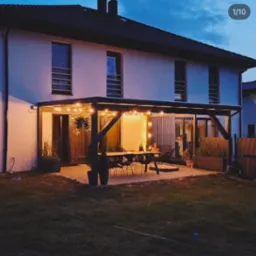 Nowoczesna pergola z ciemnego drewna, oświetlona girlandą żarówek, przylegająca do białego domu dwupiętrowego, z widocznym stołem i krzesłami na tarasie.