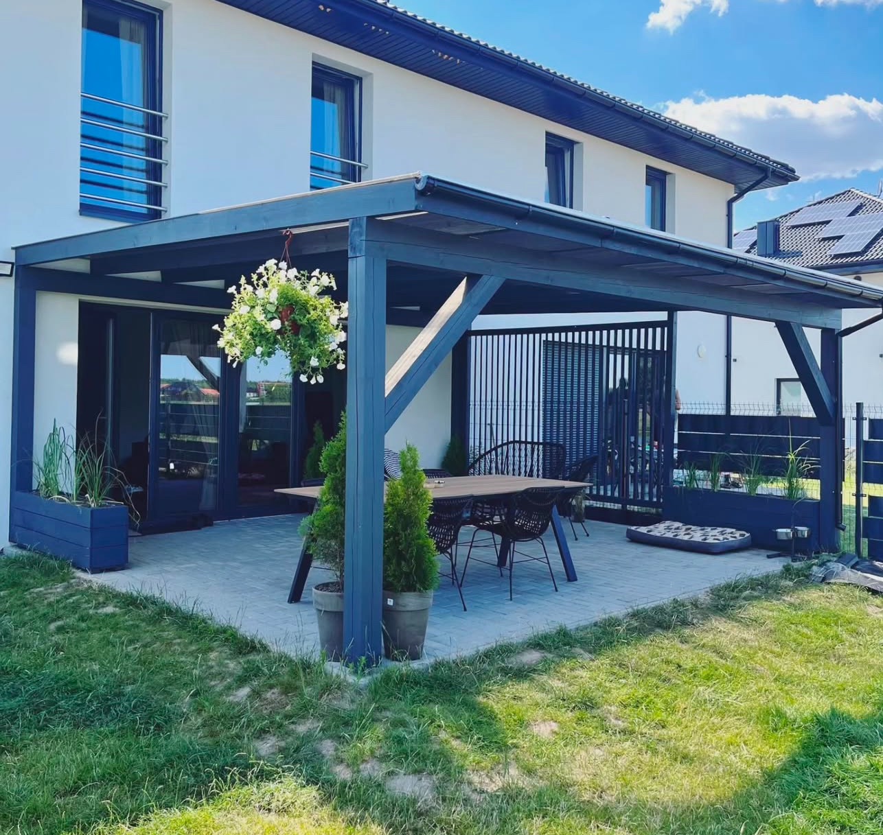 Nowoczesna, grafitowa konstrukcja zadaszenia tarasu przy białym domu, z wiszącą kompozycją kwiatową i meblami ogrodowymi, na tle błękitnego nieba.