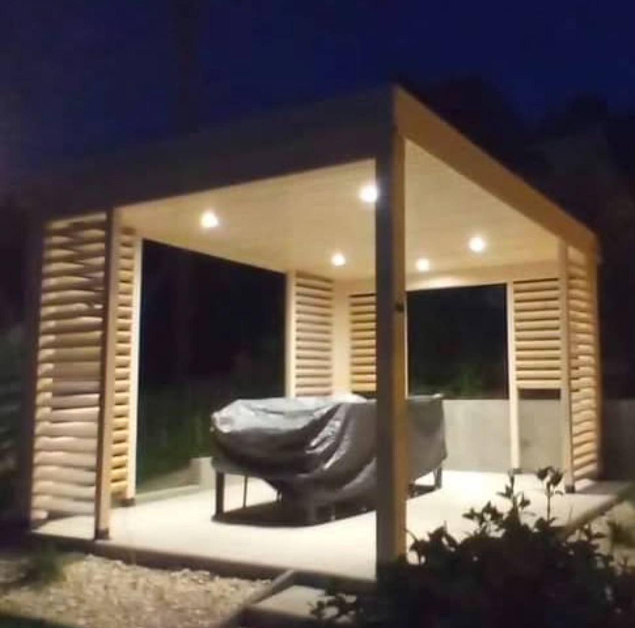 Oświetlona altana z jasnego drewna z lamelowymi ścianami bocznymi, osłonięty mebel ogrodowy wewnątrz, widok nocny.