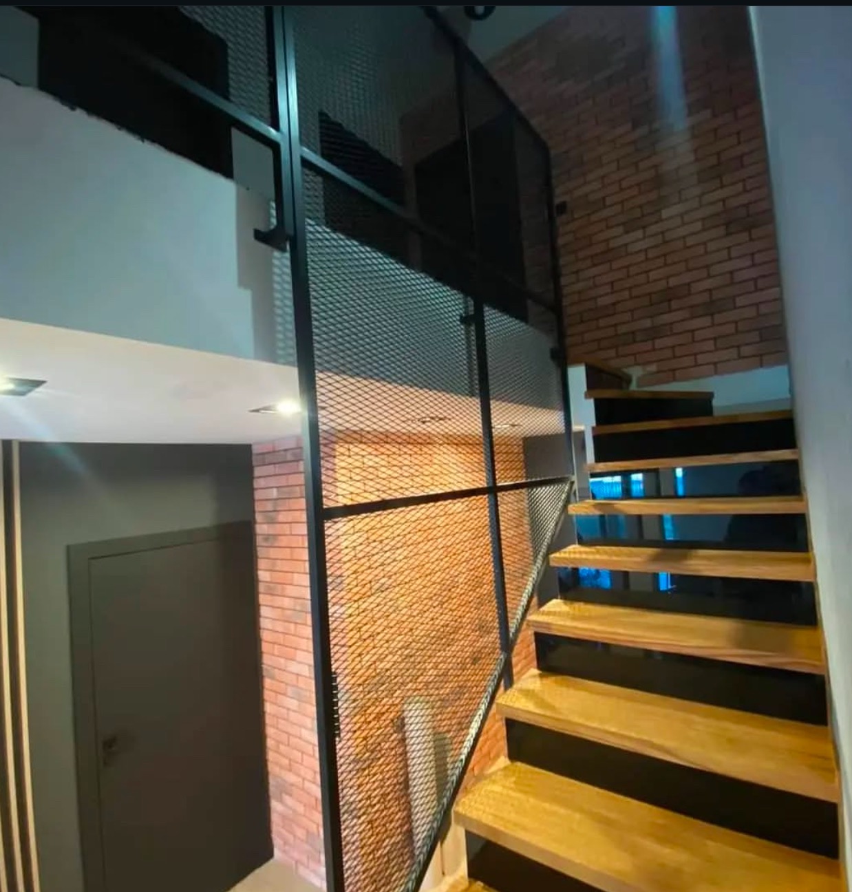 Schody drewniane z czarnymi podstopnicami i metalową balustradą z siatki, widok z dołu, ceglana ściana w tle.