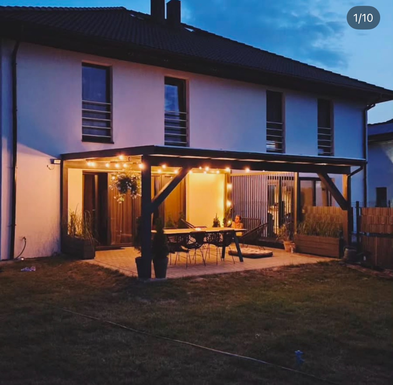 Nowoczesna pergola z ciemnego drewna, oświetlona girlandą żarówek, przylegająca do białego domu dwupiętrowego, z widocznym stołem i krzesłami na tarasie.