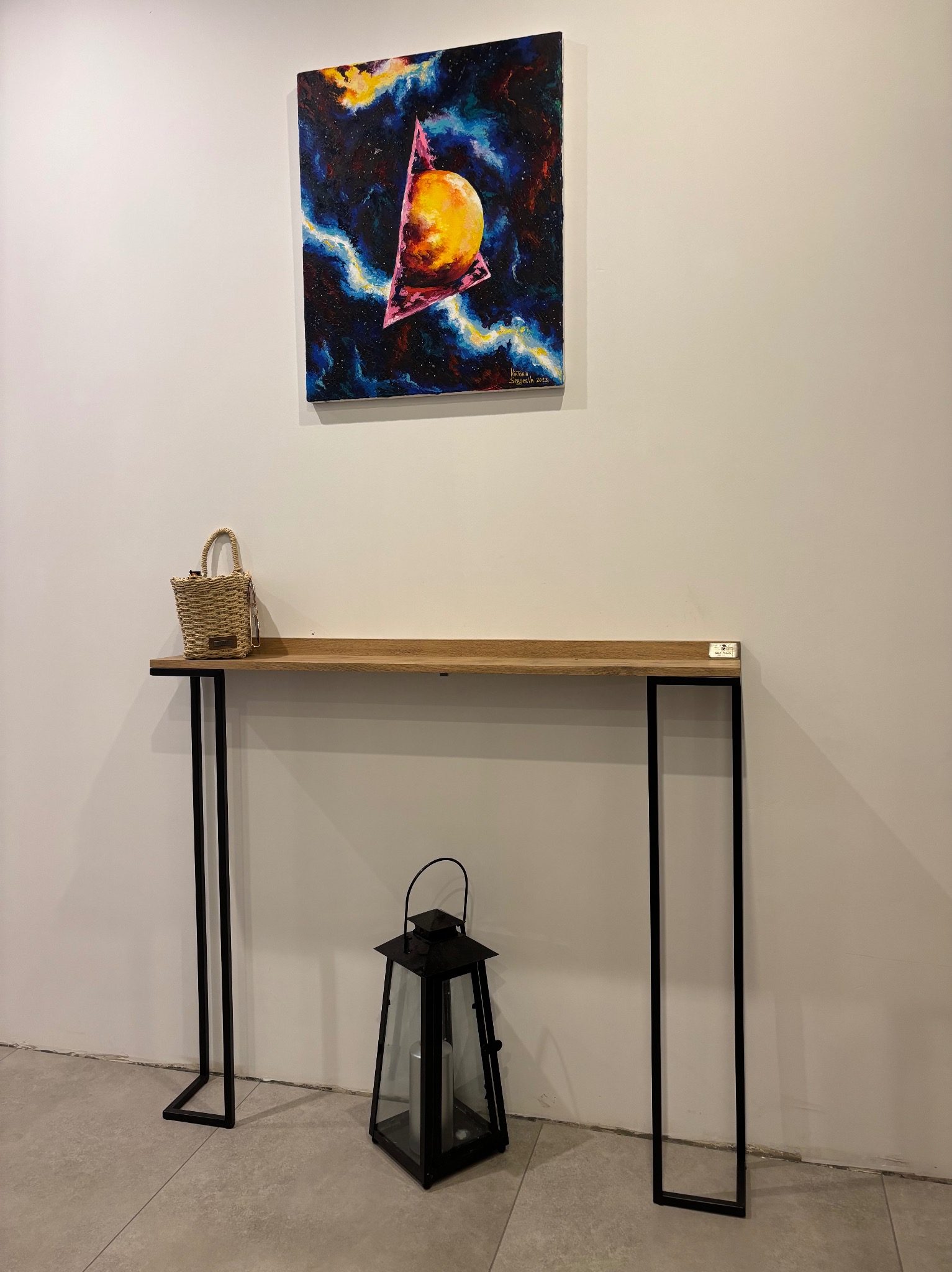 Wąska drewniana konsola na czarnych metalowych nogach, udekorowana wiklinowym koszykiem i czarnym lampionem, wiszący obraz z motywem kosmicznym w tle.
