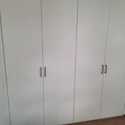 Czterodrzwiowa, biała szafa wnękowa z ciemnymi, pionowymi uchwytami na tle drewnianej podłogi.