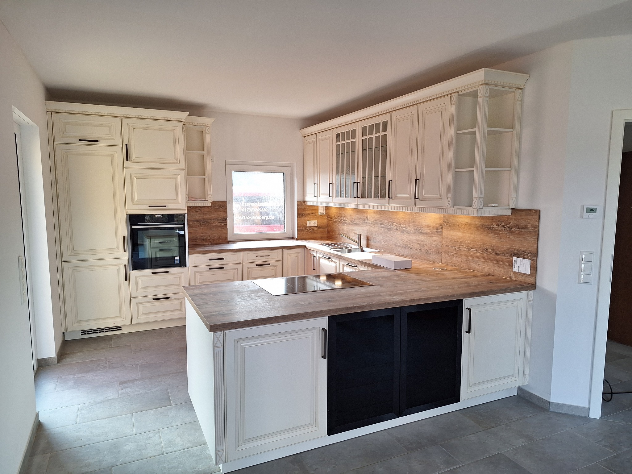 Jasna kuchnia z drewnianym blatem, wyspą z czarnymi frontami i przeszklonymi szafkami wiszącymi, oświetlona listwą LED pod szafkami.