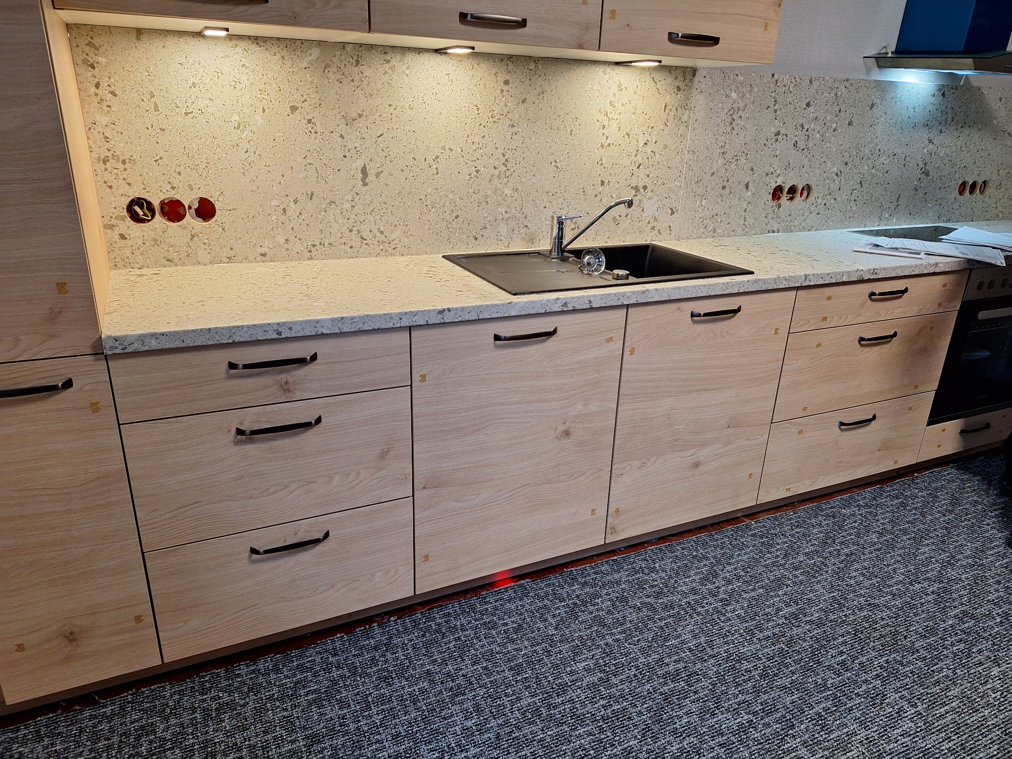 Jasna kuchnia z drewnianymi frontami, czarnym zlewem i blatem z jasnego konglomeratu. Widoczne gniazdka elektryczne w ścianie nad blatem i oświetlenie podszafkowe.