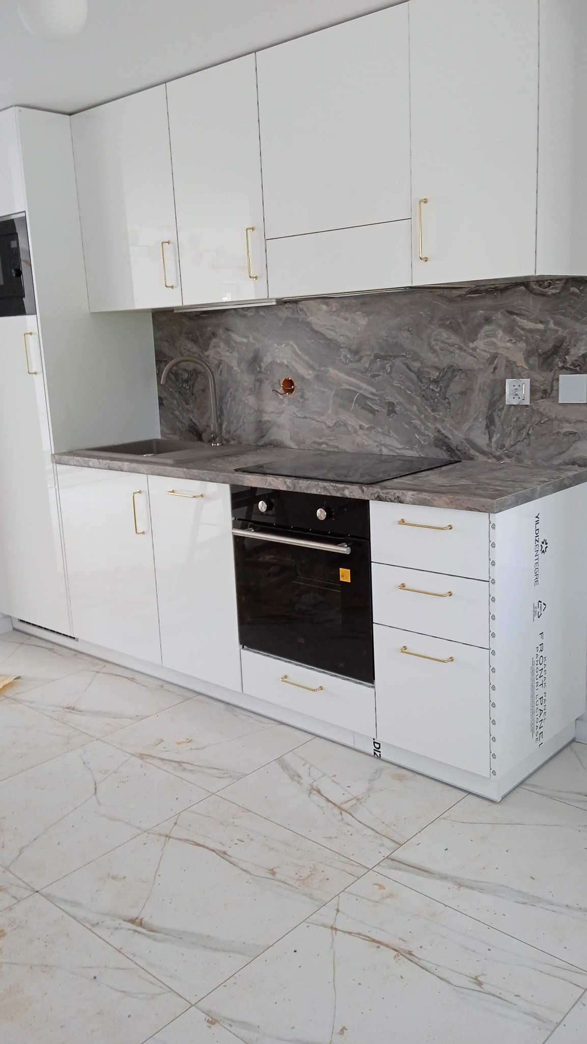 Biała, lakierowana zabudowa kuchenna z marmurowym blatem i złotymi uchwytami, widoczna płyta indukcyjna i piekarnik w zabudowie, ujęcie z niskiego kąta.