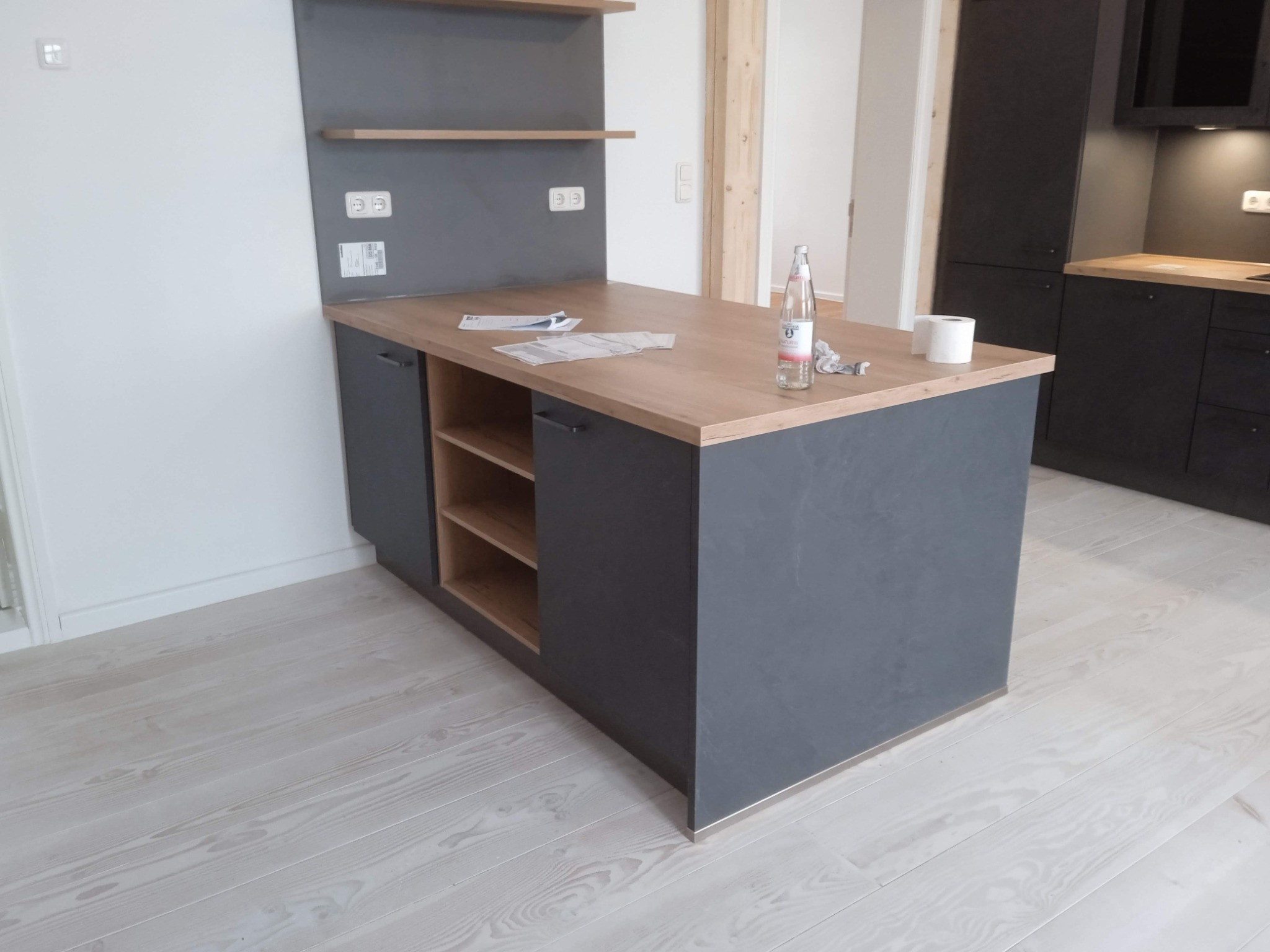 Wyspa kuchenna z szarymi frontami i drewnianym blatem, na blacie butelka wody, papier toaletowy i dokumenty, biała podłoga z desek.