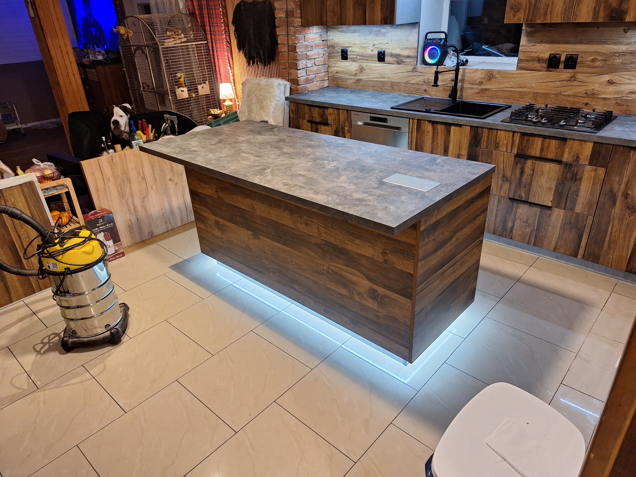 Nowoczesna wyspa kuchenna z podświetleniem LED w Banie. Blat w odcieniach szarości, fronty szafek z ciemnego drewna. Widoczna zabudowa kuchenna w tle.