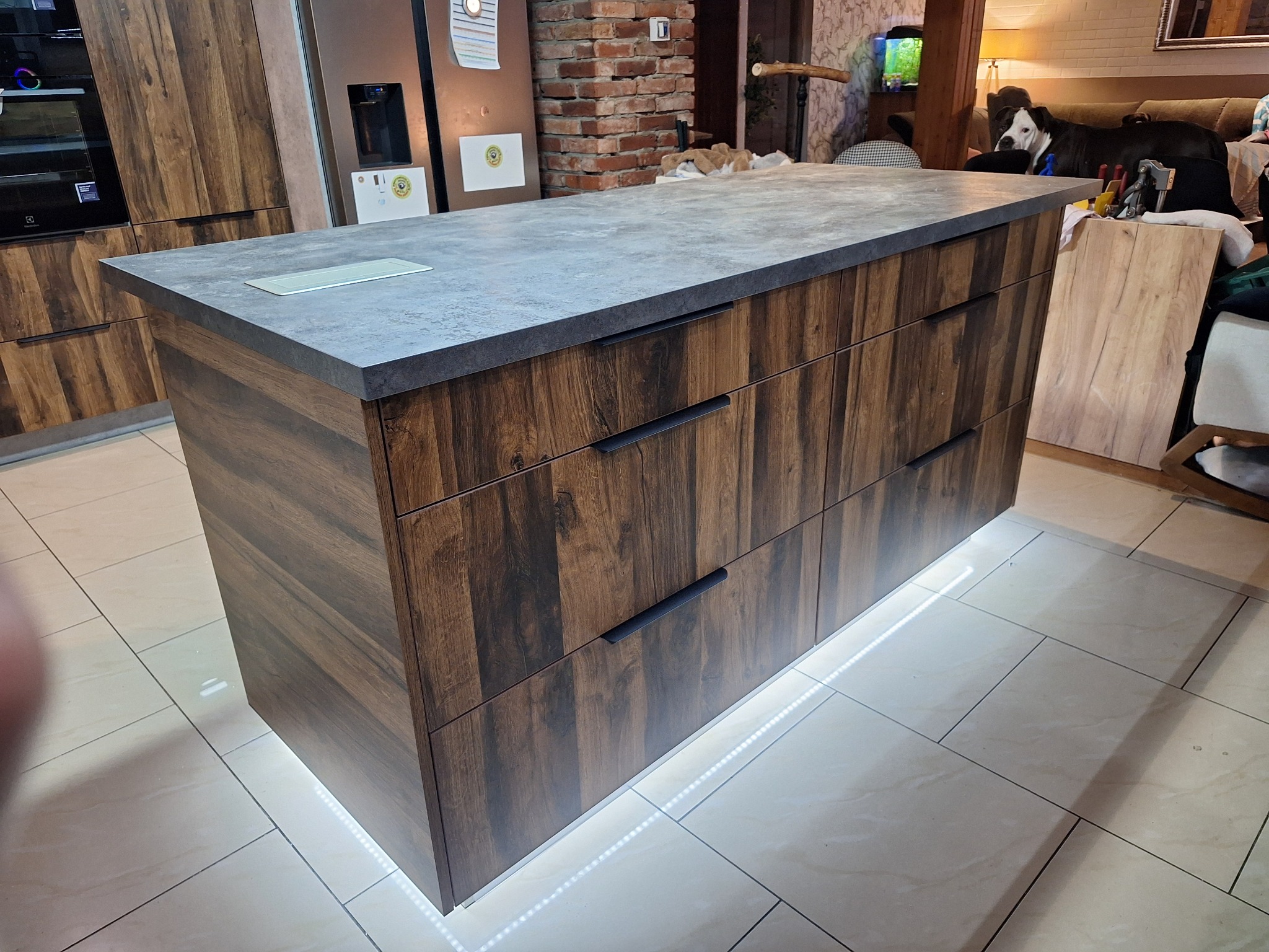 Wyspa kuchenna z szufladami w odcieniach brązu i szarym blatem, podświetlona od dołu. W tle widoczna zabudowa kuchenna i ceglana ściana.