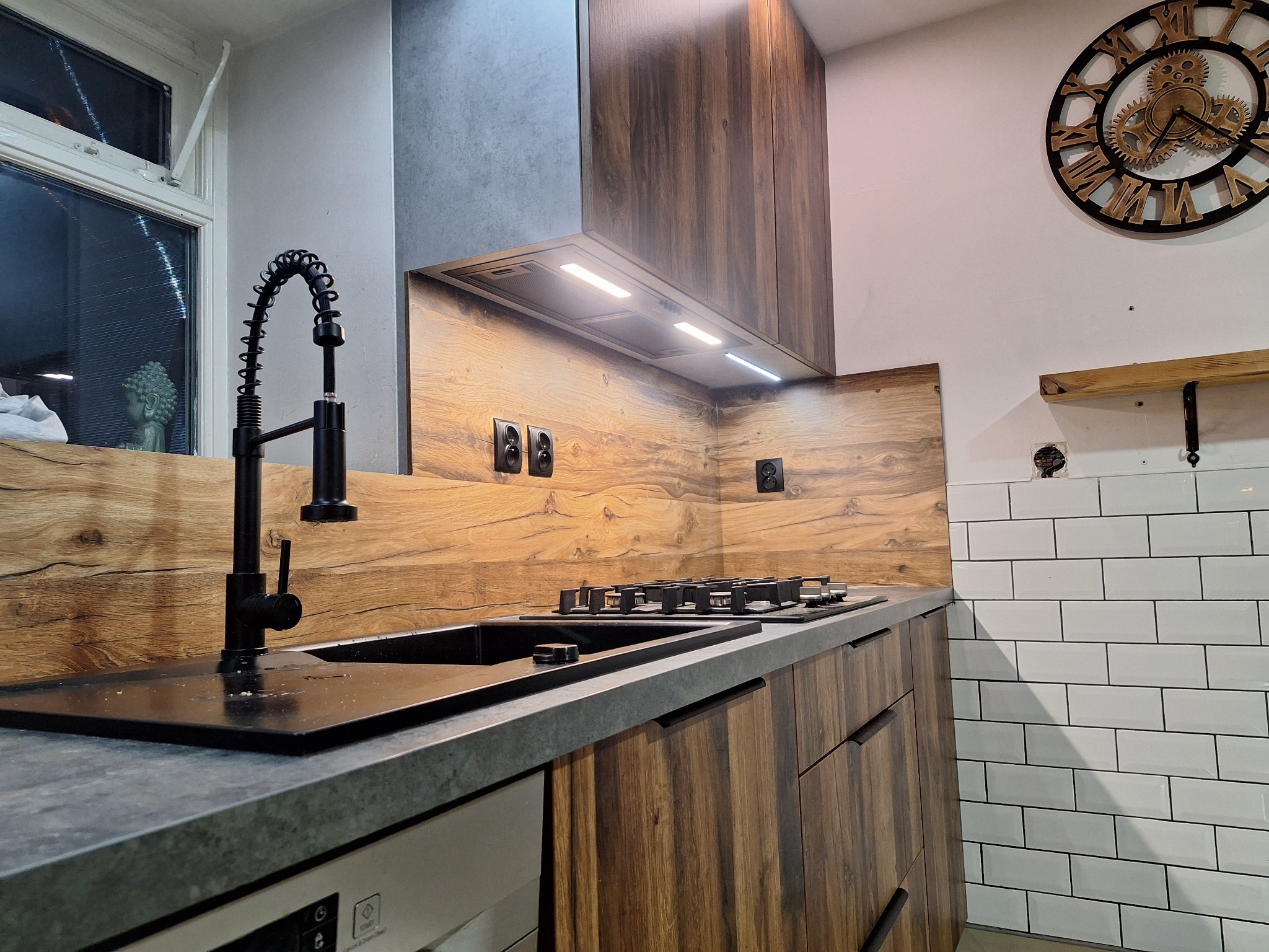 Nowoczesna kuchnia z drewnianymi frontami, szarym blatem i czarną armaturą. Na ścianie dekoracyjny zegar i białe płytki. Widoczny fragment okna z figurką Buddy.