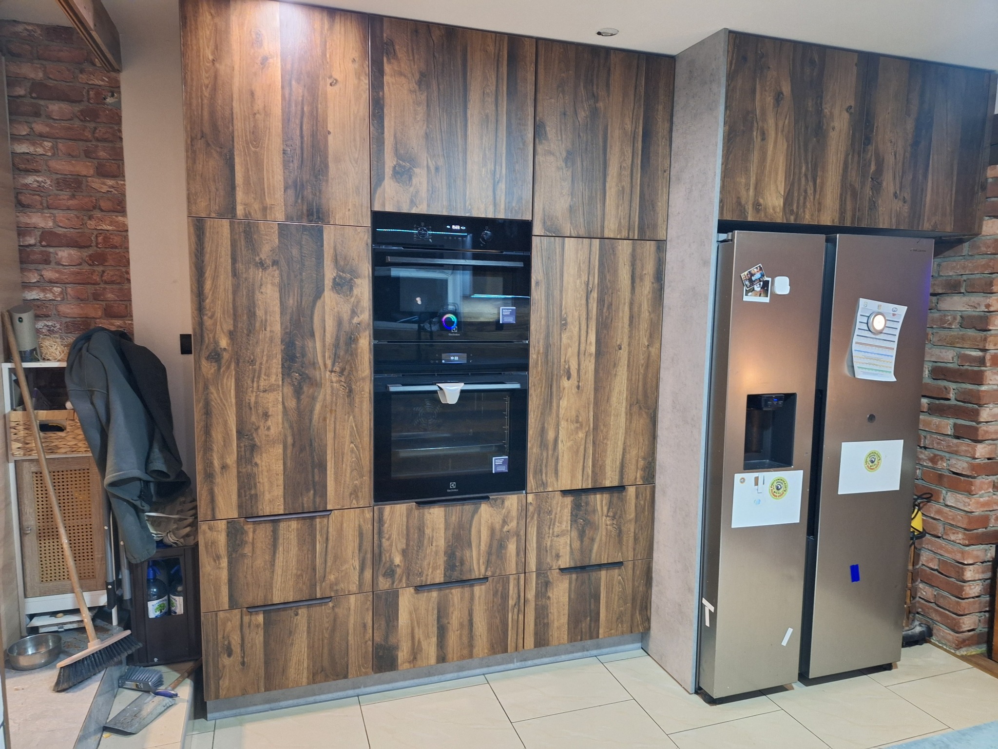 Kuchnia z frontami z ciemnego drewna, zabudowana lodówka side-by-side, piekarnik i mikrofalówka w zabudowie. Ceglana ściana w tle dodaje charakteru.