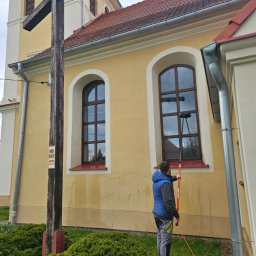 Czysta Sprawa 24 CLEAN CASE Sp. z o.o. - Pracownik w niebieskiej kamizelce myje wysokie okna kościoła za pomocą teleskopowego kija z myjką; widoczne zacieki na elewacji.