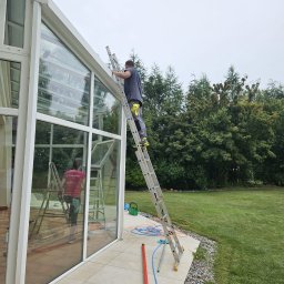 Czysta Sprawa 24 CLEAN CASE Sp. z o.o. - Pracownik na drabinie myje okna oranżerii, widoczne narzędzia i drugi pracownik wewnątrz, na zewnątrz trawnik i drzewa.