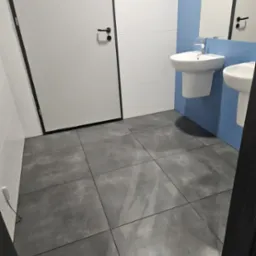 Wnętrze łazienki z dwoma umywalkami na tle niebieskich płytek, szare płytki podłogowe, białe ściany i drzwi z czarnymi obramowaniami.