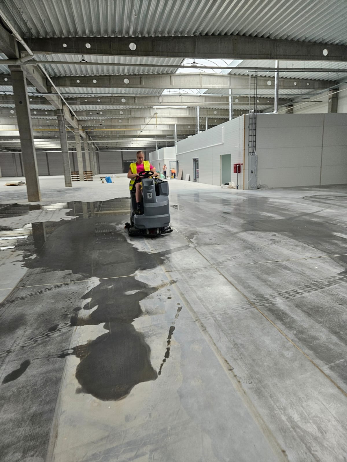 Pracownik w żółtej kamizelce odblaskowej obsługuje szorowarkę na mokrej posadzce w hali przemysłowej, widoczne ślady opon i plamy wody.