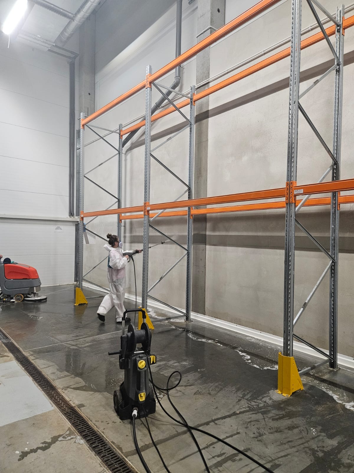 Pracownik w kombinezonie ochronnym myje ścianę magazynu pod regałami za pomocą myjki ciśnieniowej, obok widoczna maszyna czyszcząca podłogę.