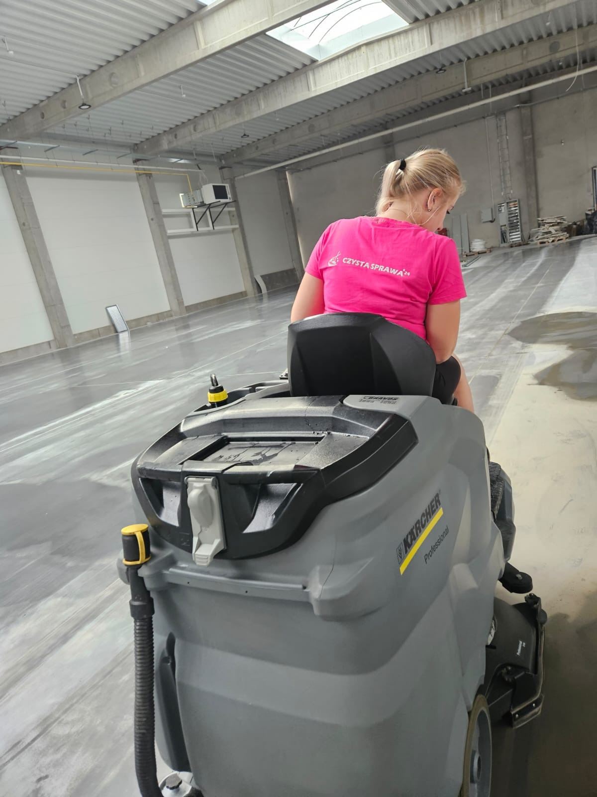 Pracownica w różowej koszulce z napisem 'Czysta Sprawa' na maszynie czyszczącej Karcher Professional, myjąca posadzkę w hali przemysłowej z betonowym stropem i świetlikami.