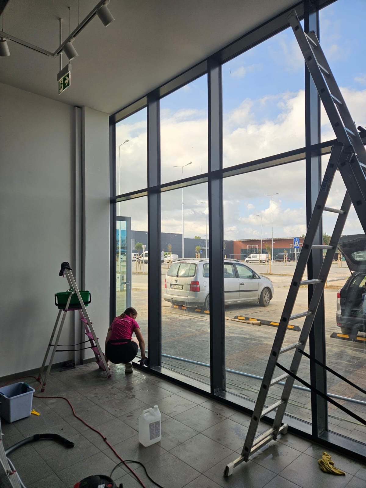 Kobieta w różowej koszulce czyści dolną krawędź dużej, przeszklonej ściany w biurze, używając ściereczki; widoczne dwie drabiny, wiadro, odkurzacz i płyn do czyszczenia, a za szybą parking...