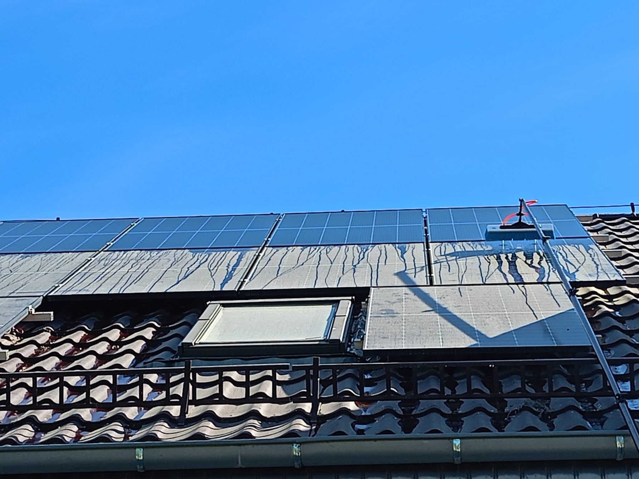 Czyszczenie paneli słonecznych na dachu domu z użyciem teleskopowej szczotki, widoczne zacieki wody na panelach i dachówce.