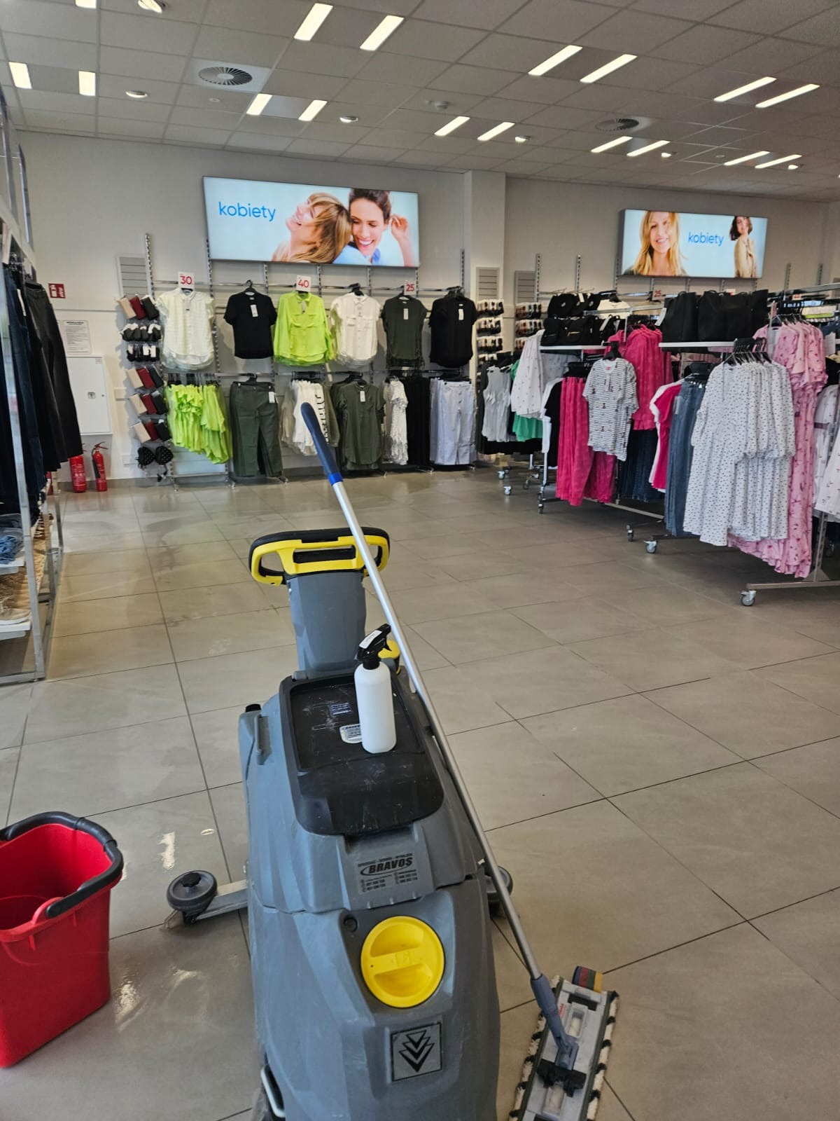 Automat do mycia podłóg marki Bravos z mopem na jasnych płytkach w sklepie odzieżowym, w tle wieszaki z ubraniami i reklamy świetlne.