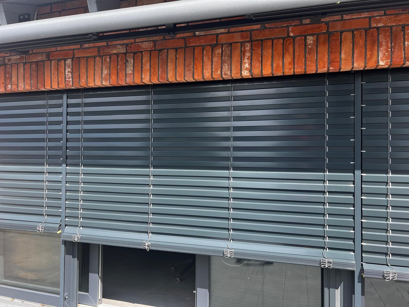 Zewnętrzne, szare żaluzje aluminiowe zamontowane na oknach budynku z ceglaną elewacją, częściowo opuszczone, widoczne sprężynowe zaczepy.