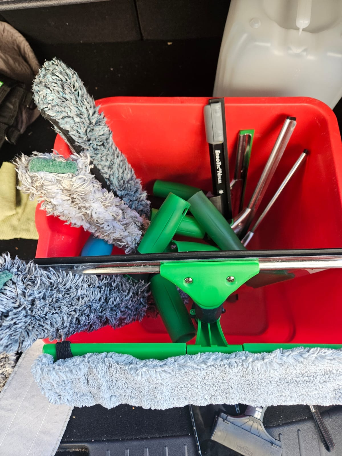 Czerwone wiadro wypełnione narzędziami do mycia okien: ściągaczki z zielonymi uchwytami, myjki z szarym włóknem i inne akcesoria.