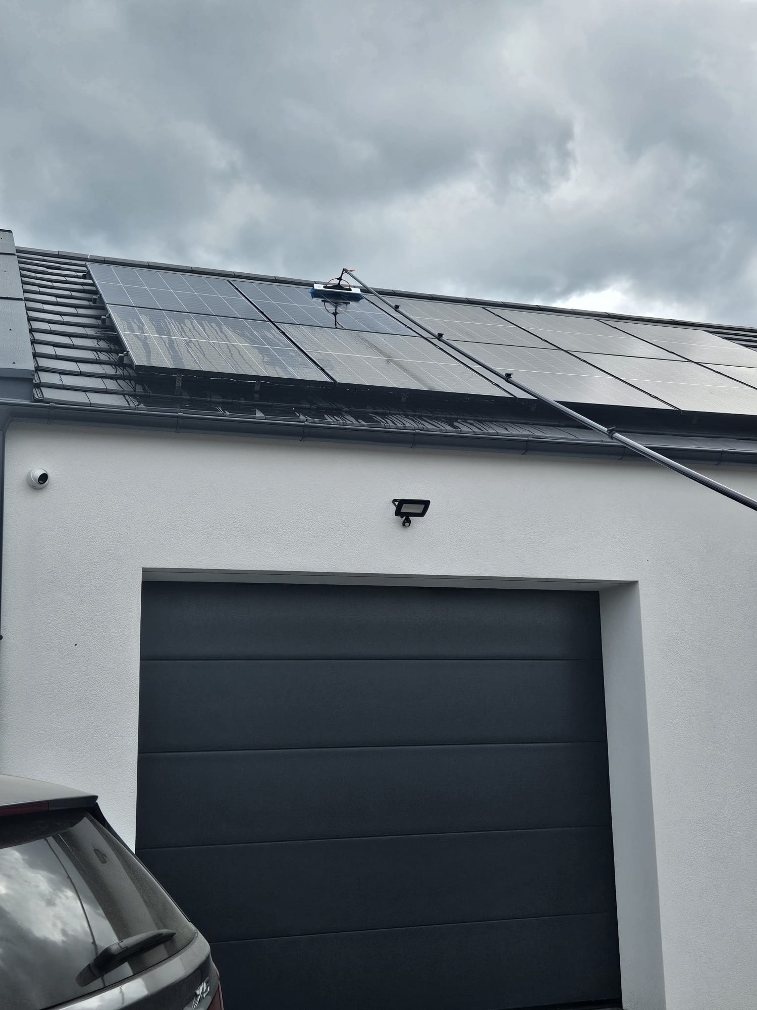 Czyszczenie paneli słonecznych na dachu domu za pomocą szczotki na długim wysięgniku, widoczne ślady brudu i wody na panelach, pochmurne niebo w tle.