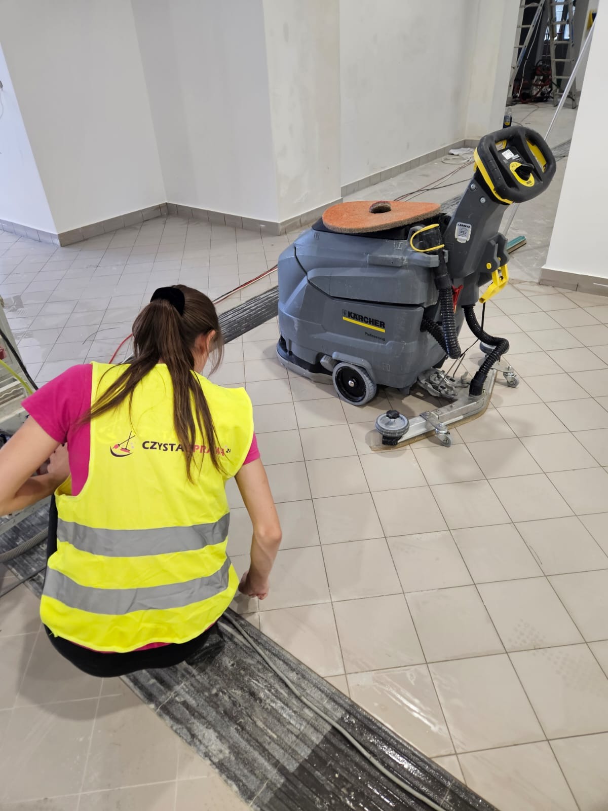 Pracownica w żółtej kamizelce czyści podłogę z płytek za pomocą szorowarki Karcher, widoczny brud i woda na czyszczonej powierzchni, w tle fragment drabiny.