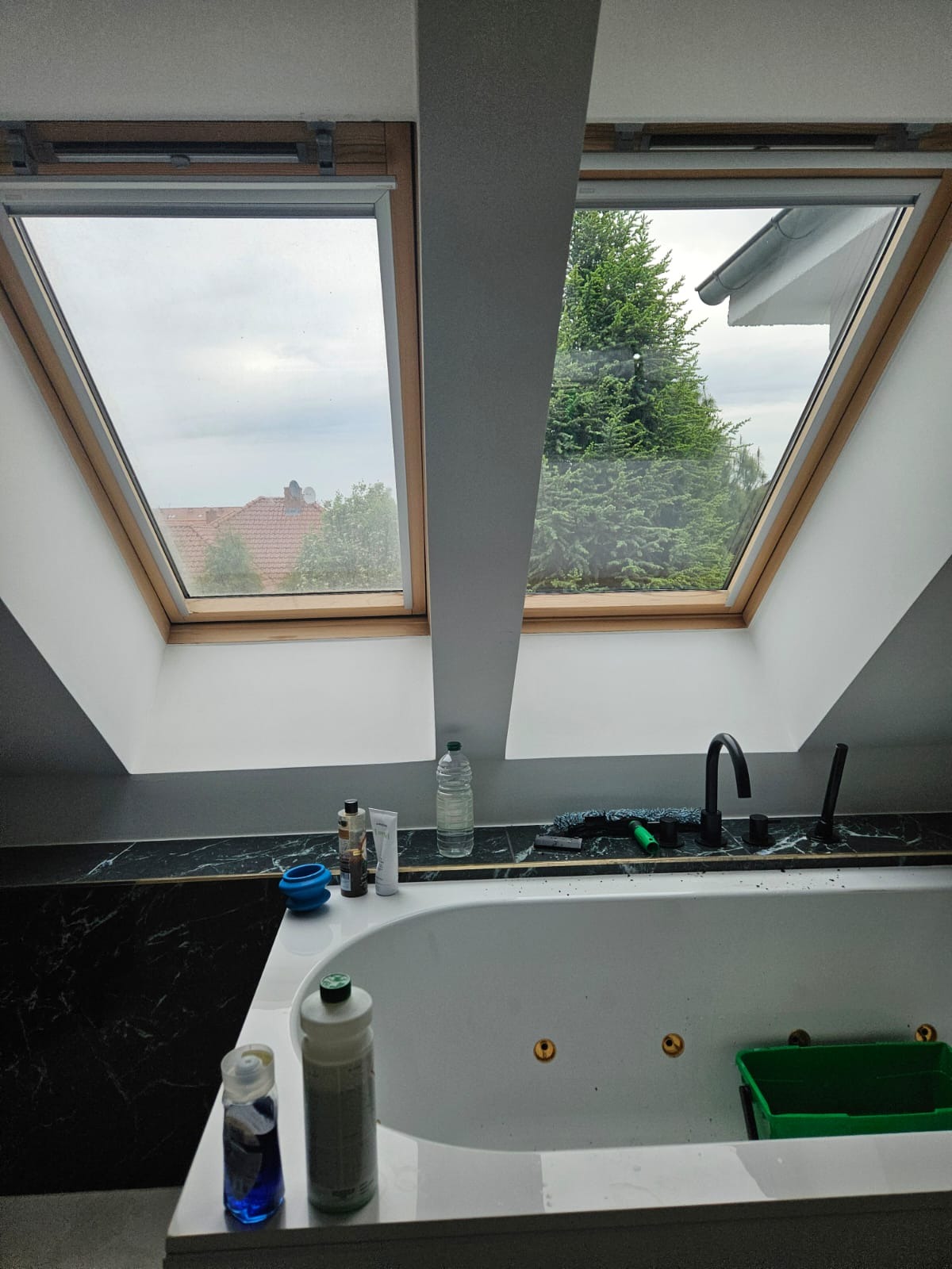 Dwa okna dachowe z widokiem na zewnątrz, wanna z hydromasażem i akcesoria do czyszczenia w łazience na poddaszu.