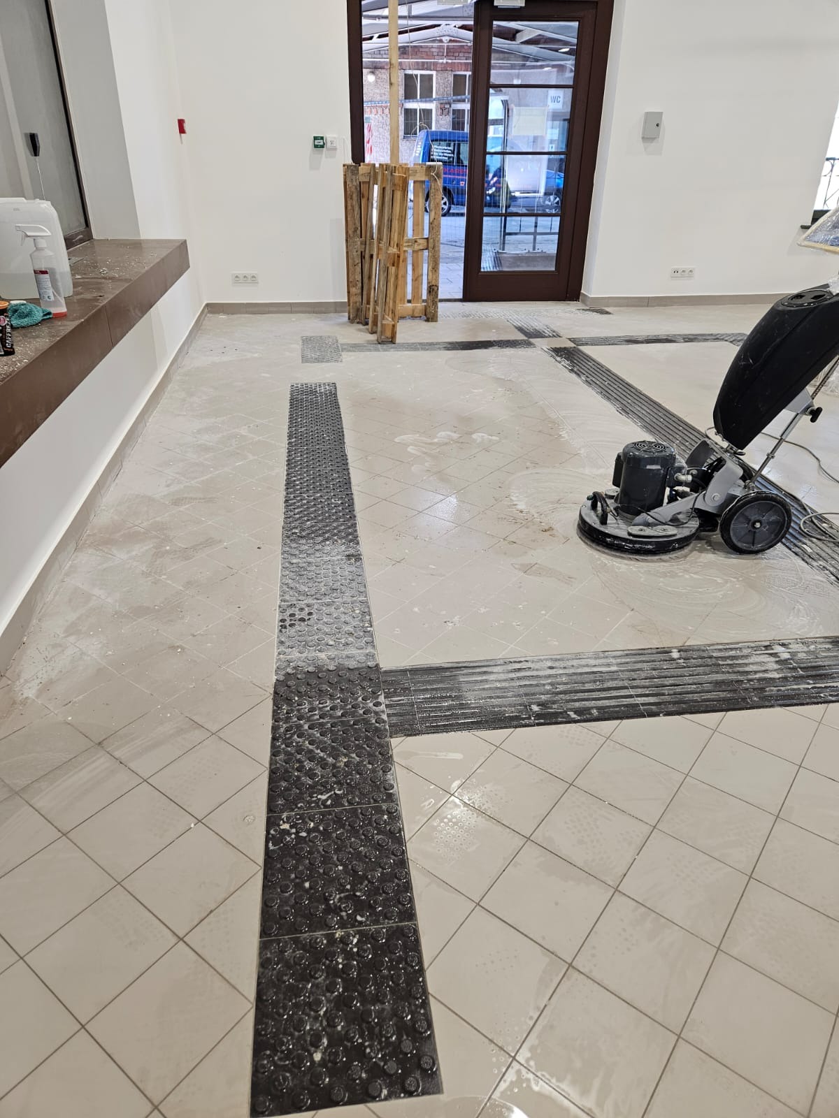 Maszyna czyszcząca podłogę w trakcie pracy w przestronnym, jasnym pomieszczeniu z płytkami i elementami dla osób niedowidzących, widoczne ślady po czyszczeniu i resztki detergentu.