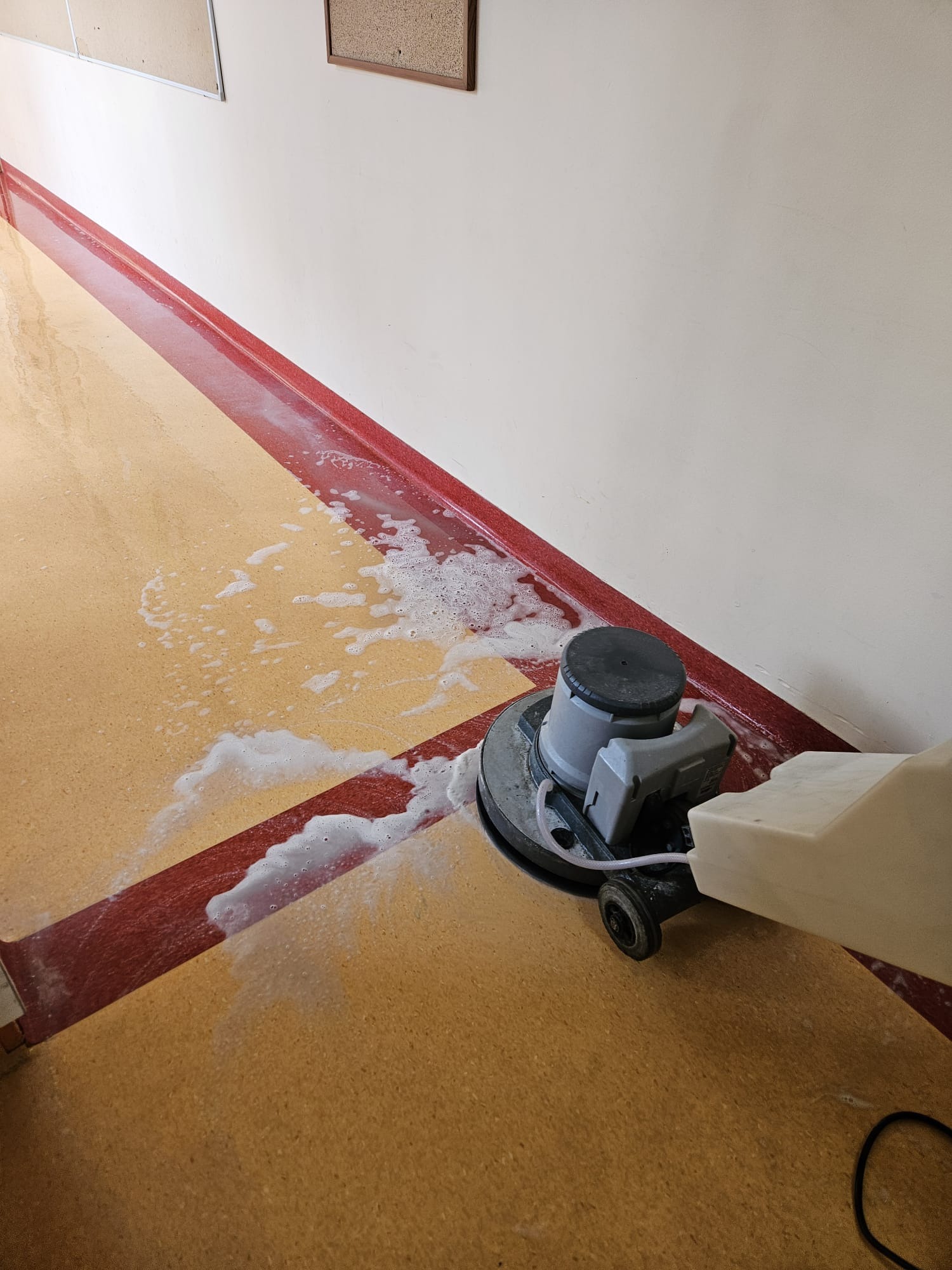 Maszyna do czyszczenia podłóg w trakcie pracy, widoczne ślady piany i wilgoci na powierzchni, czerwony pas przy ścianie.