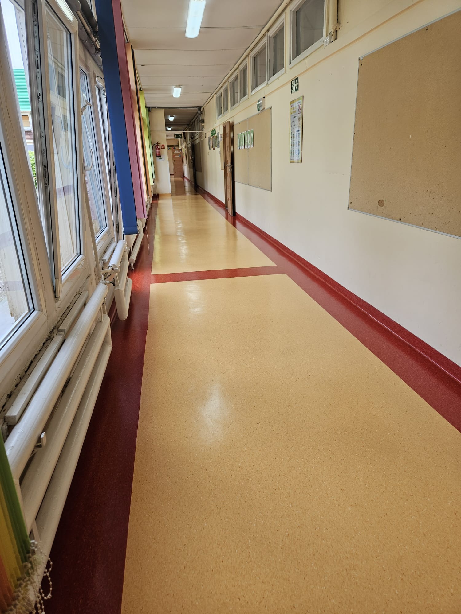 Długi korytarz w budynku użyteczności publicznej z otwartymi oknami po lewej stronie, tablicami ogłoszeń na ścianie po prawej i linoleum na podłodze.
