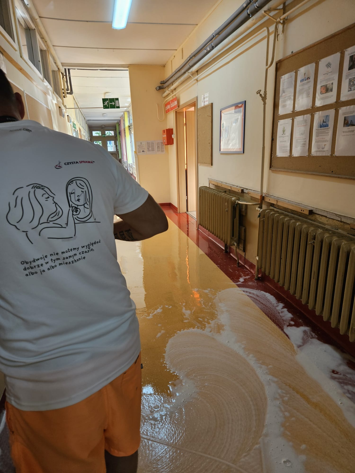 Pracownik w koszulce z napisem 'Czysta Sprawa' czyści podłogę w korytarzu budynku w Gralewie; widoczne ślady piany i mokra powierzchnia, grzejniki przy ścianie.